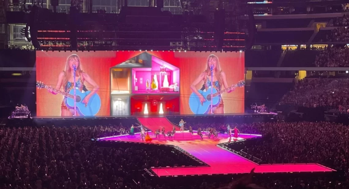 A female pop singer performs with a guitar on a brightly lit stadium stage, with giant screens behind her, dancers on the runway, and a massive crowd filling the arena.