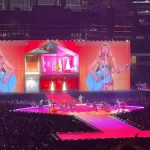 A female pop singer performs with a guitar on a brightly lit stadium stage, with giant screens behind her, dancers on the runway, and a massive crowd filling the arena.