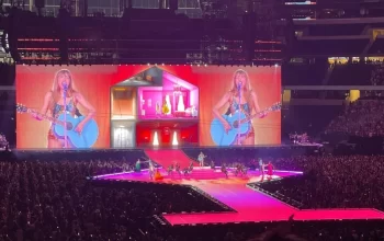 A female pop singer performs with a guitar on a brightly lit stadium stage, with giant screens behind her, dancers on the runway, and a massive crowd filling the arena.