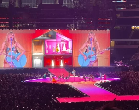 A female pop singer performs with a guitar on a brightly lit stadium stage, with giant screens behind her, dancers on the runway, and a massive crowd filling the arena.