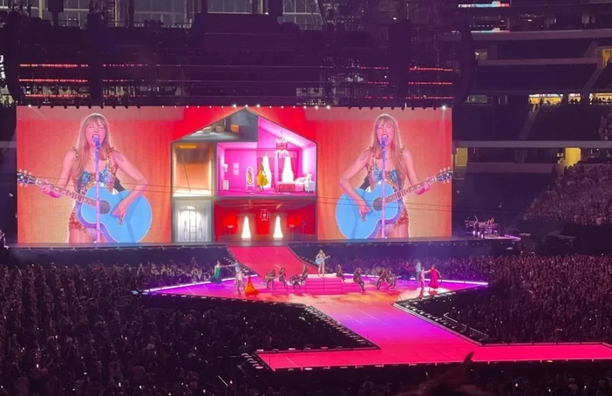 A female pop singer performs with a guitar on a brightly lit stadium stage, with giant screens behind her, dancers on the runway, and a massive crowd filling the arena.