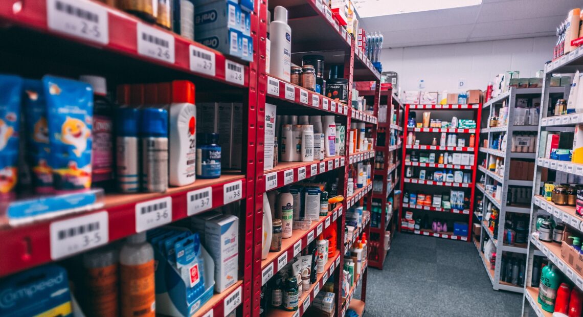 Long aisles of a retail stockroom with red and white metal shelves filled with boxed and bottled consumer products, including personal care items, arranged with barcode labels under bright overhead lighting.