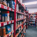 Long aisles of a retail stockroom with red and white metal shelves filled with boxed and bottled consumer products, including personal care items, arranged with barcode labels under bright overhead lighting.