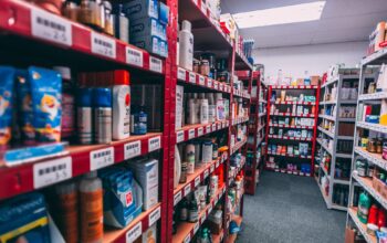 Long aisles of a retail stockroom with red and white metal shelves filled with boxed and bottled consumer products, including personal care items, arranged with barcode labels under bright overhead lighting.