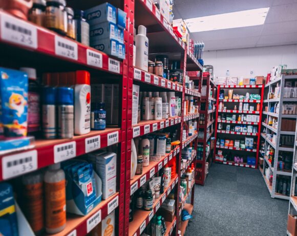 Long aisles of a retail stockroom with red and white metal shelves filled with boxed and bottled consumer products, including personal care items, arranged with barcode labels under bright overhead lighting.
