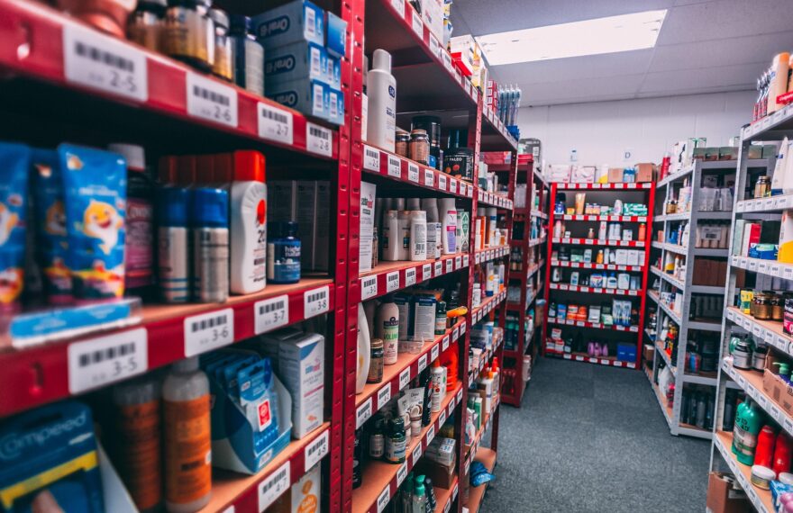 Long aisles of a retail stockroom with red and white metal shelves filled with boxed and bottled consumer products, including personal care items, arranged with barcode labels under bright overhead lighting.