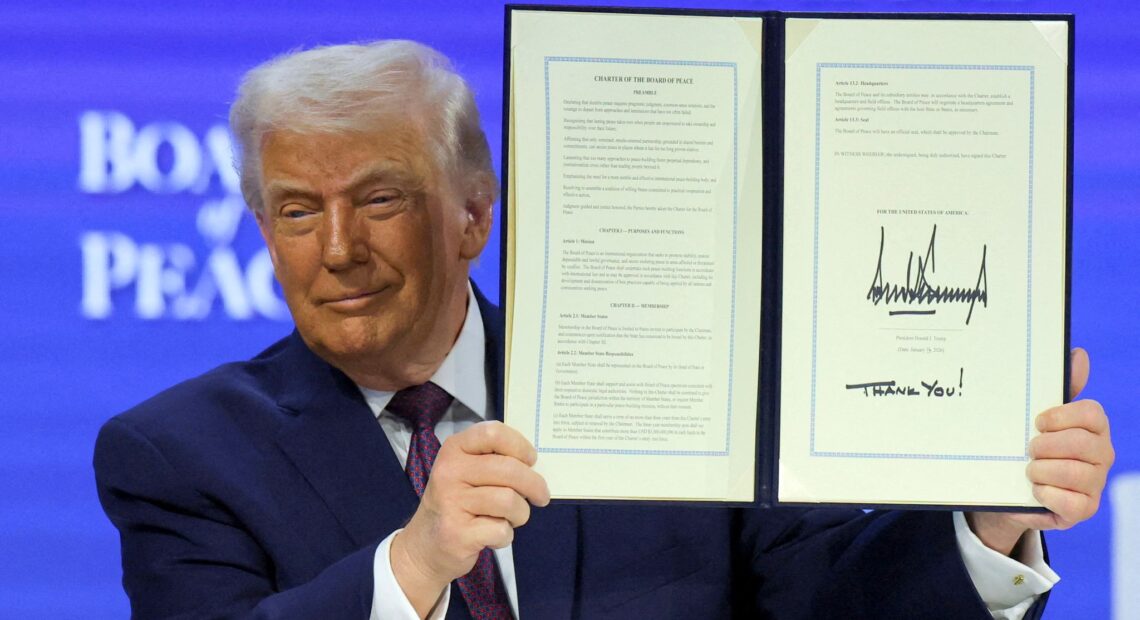 A man in a dark suit holds up an open, signed document titled “Charter of the Board of Peace,” displaying two framed pages against a blue conference backdrop.