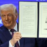 A man in a dark suit holds up an open, signed document titled “Charter of the Board of Peace,” displaying two framed pages against a blue conference backdrop.