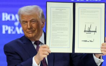 A man in a dark suit holds up an open, signed document titled “Charter of the Board of Peace,” displaying two framed pages against a blue conference backdrop.