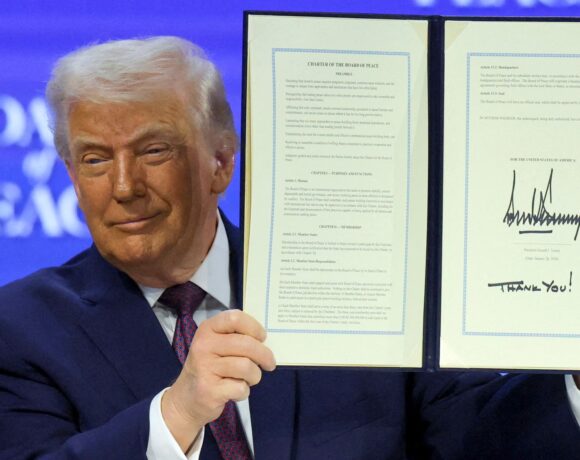 A man in a dark suit holds up an open, signed document titled “Charter of the Board of Peace,” displaying two framed pages against a blue conference backdrop.