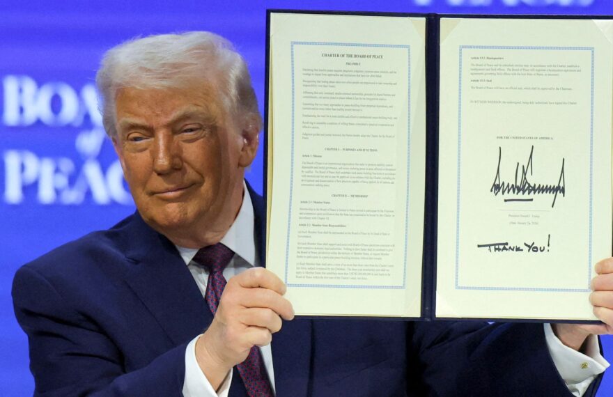 A man in a dark suit holds up an open, signed document titled “Charter of the Board of Peace,” displaying two framed pages against a blue conference backdrop.